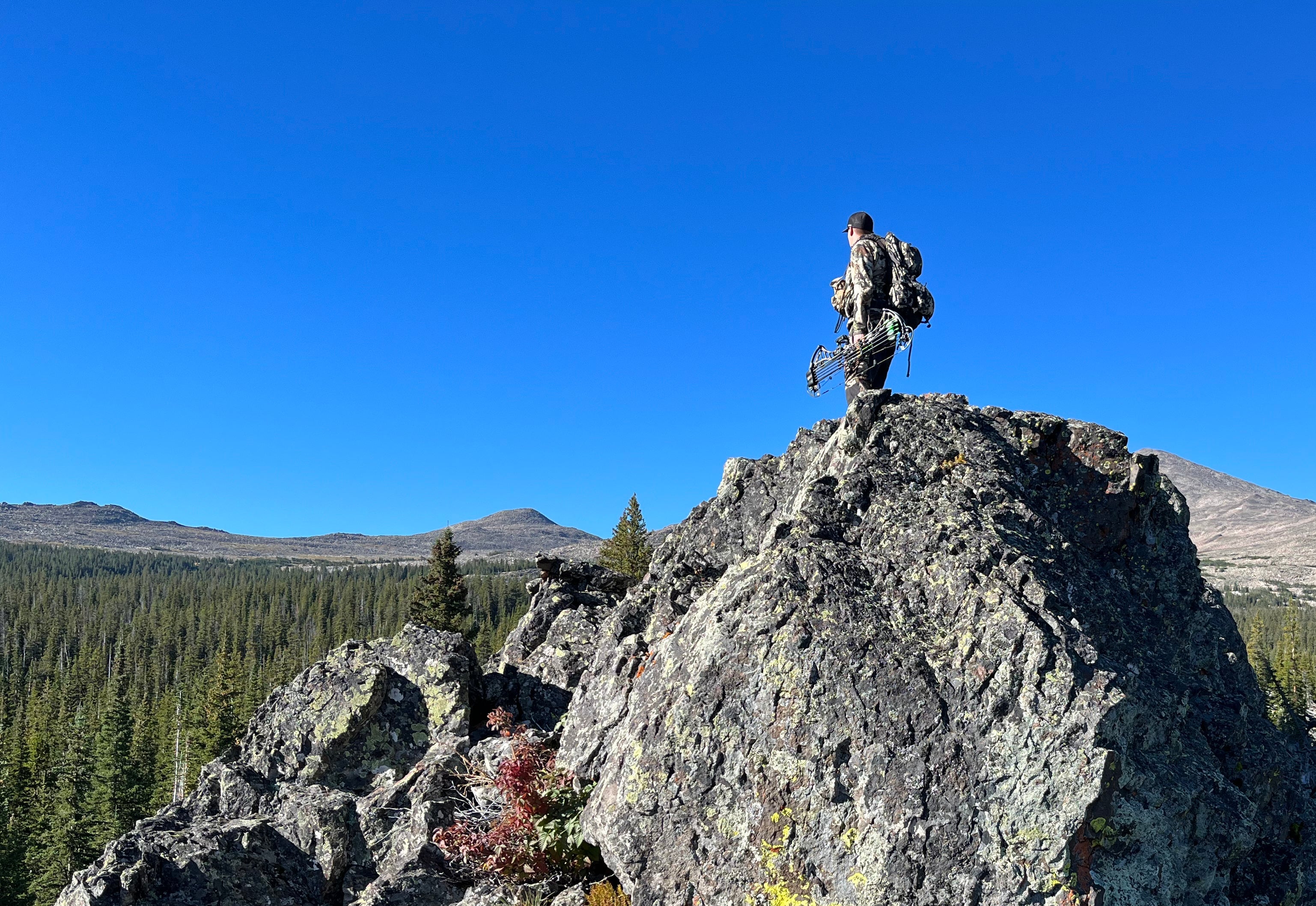 hunter with compound bow standing on a rocky outcrop with a clear blue sky and forested landscape in the background